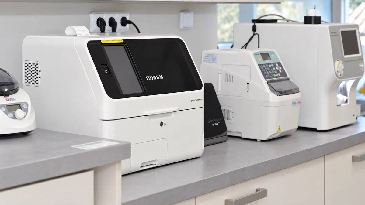 Šikmý pohled na řadu analyzátorů krevních vzorků na pultu laboratoře, v popředí biochemický analyzátor Fujifilm