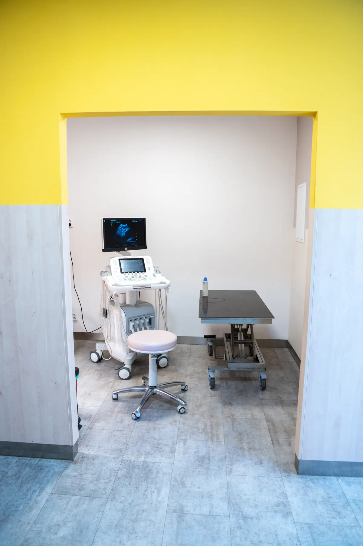 Pohled otevřenými dveřmi do místnosti pro ultrazvuk, kde je otočná stolička; vidět je také žlutá stěna
